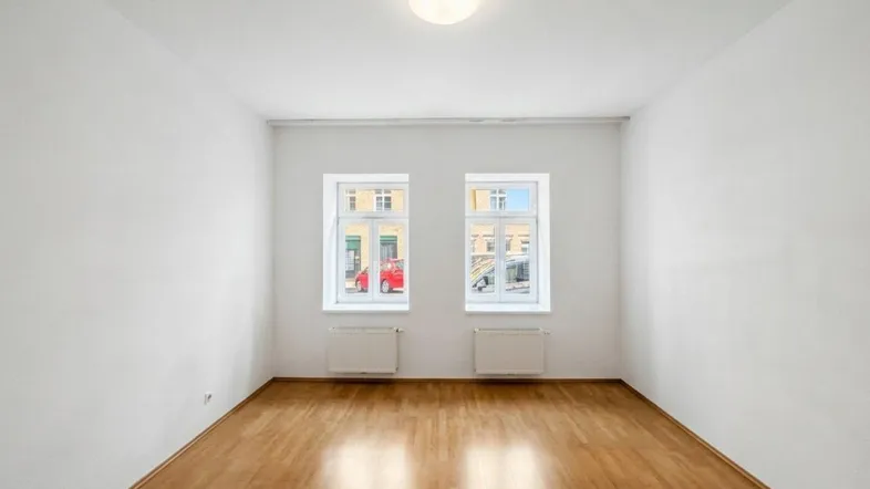 Helles, leeres Zimmer mit Parkettboden und zwei Fenstern, bereit zur individuellen Gestaltung.
