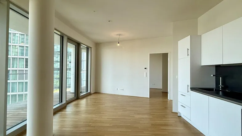 Heller offener Wohnbereich mit integrierter Küche, großen Fenstern und Zugang zum Balkon.