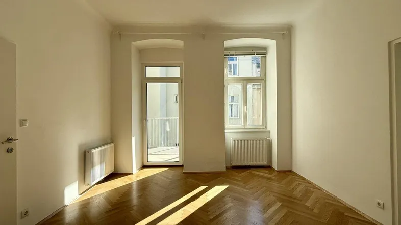 Helles Zimmer mit Fischgrätparkett, weißen Wänden und Zugang zu einem Balkon, durchflutet von Sonnenlicht.