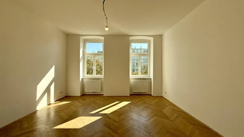 Helles Zimmer mit Fischgrätparkettboden und zwei Fenstern, die viel Sonnenlicht hereinlassen.