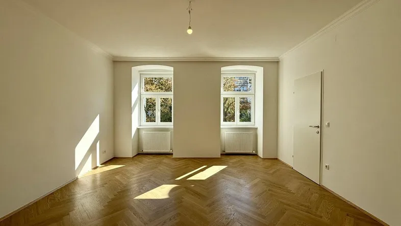 Helles, leeres Zimmer mit Fischgrätparkettboden, hohen Decken und zwei Fenstern, die viel Tageslicht hereinlassen.
