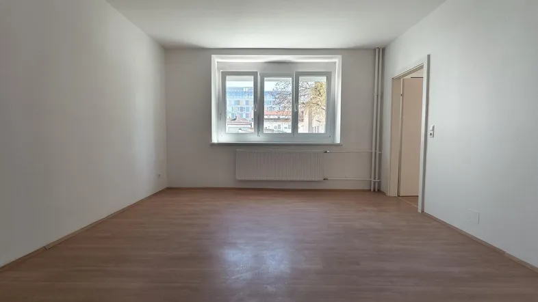 Helles, leeres Zimmer mit Laminatboden, weißen Wänden und einem Fenster, bereit zur individuellen Gestaltung.