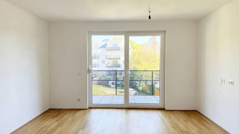 Helles Zimmer mit Parkettboden und Zugang zu einem kleinen Balkon mit Blick ins Grüne.