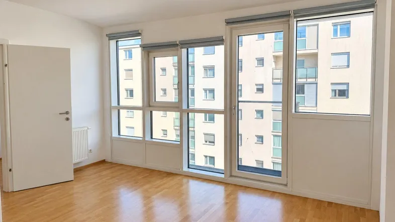 Helles Zimmer mit Parkettboden und großen Fenstern, die einen Blick auf das gegenüberliegende Gebäude bieten.