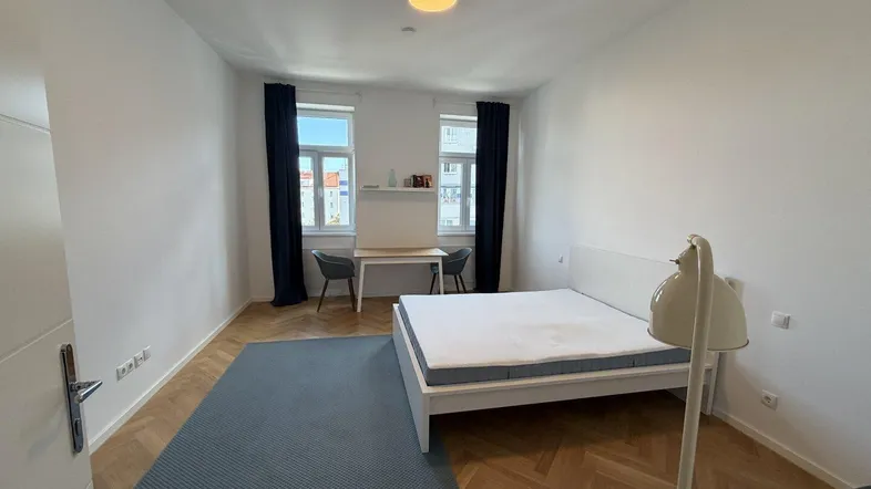 Helles Schlafzimmer mit Doppelbett, Parkettboden und zwei Fenstern mit dunklen Vorhängen.
