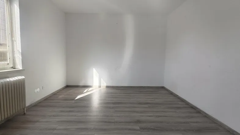 Helles, leeres Zimmer mit Laminatboden und Heizkörper unter dem Fenster.