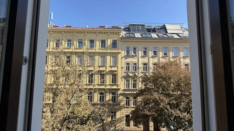 Blick aus dem Fenster auf gegenüberliegende Gebäude und Bäume unter blauem Himmel.