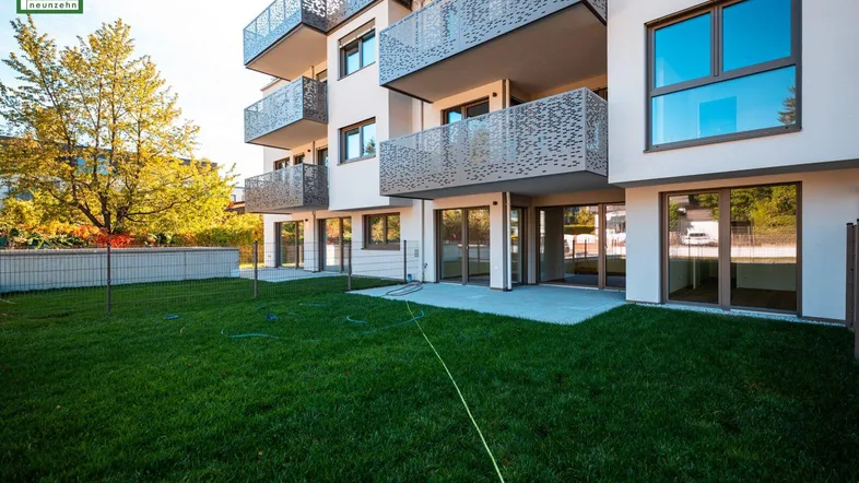 Moderner Neubau mit gepflegtem Rasen und Terrassen im Erdgeschoss, umgeben von grüner Landschaft.