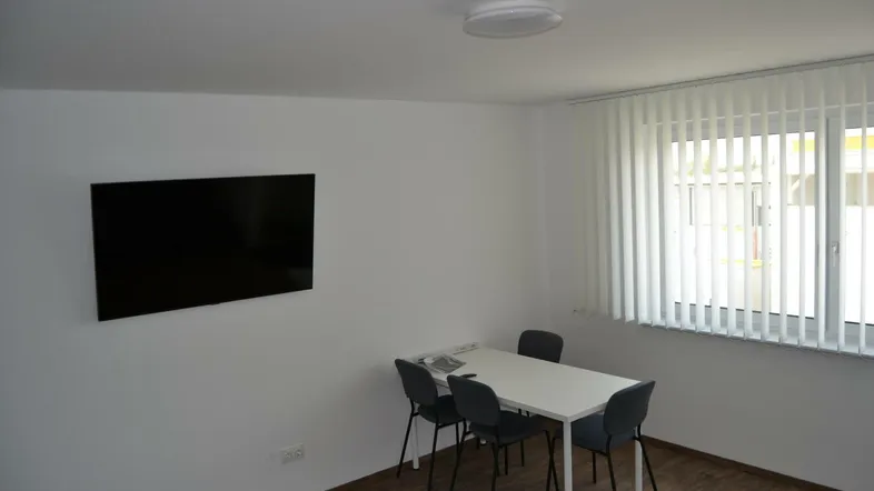 Heller Raum mit einem Tisch, vier Stühlen und einem Wandfernseher, ideal als Büro oder Besprechungszimmer.