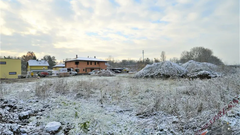 Unbebautes Grundstück mit leichter Schneedecke und Bauaktivitäten im Hintergrund unter bewölktem Himmel.