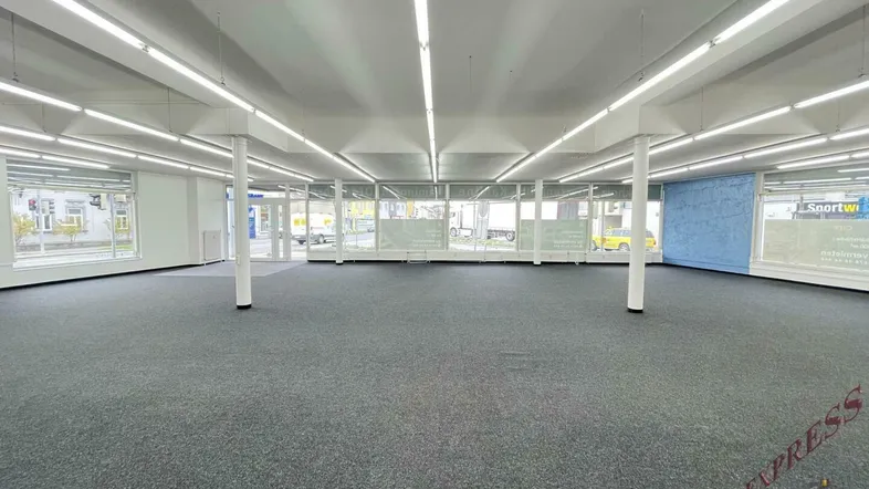 Großer, leerer Verkaufsraum mit Teppichboden und vielen Fenstern zur Straße, ideal für Einzelhandel.
