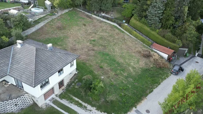 Luftaufnahme eines großen unbebauten Grundstücks neben einem Einfamilienhaus mit Satteldach und grüner Umgebung.