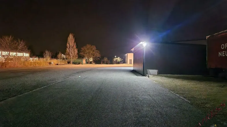 Nachtaufnahme eines großen Schotterparkplatzes mit beleuchteten Gebäuden im Hintergrund und dunklem Himmel.