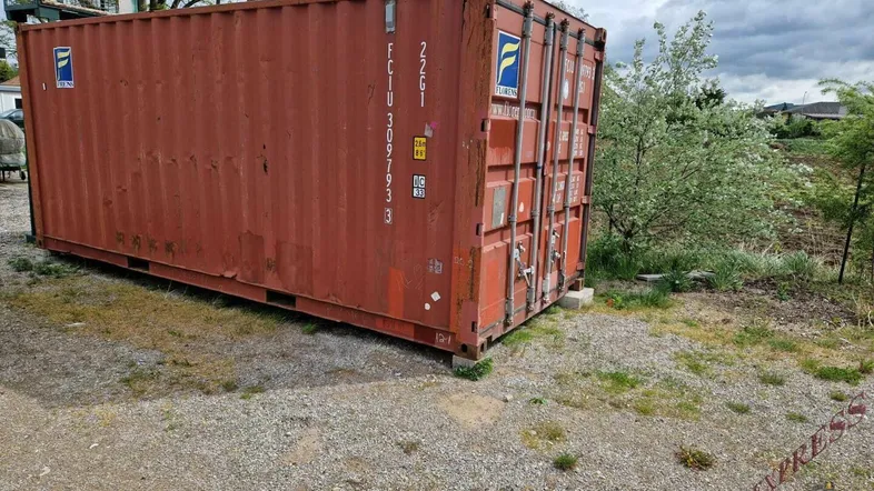 Rostiger roter Schiffscontainer auf einem Schotterplatz unter bewölktem Himmel, umgeben von spärlicher Vegetation.