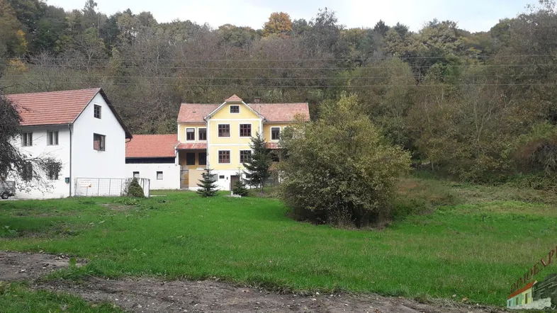 Zwei traditionelle Häuser in ländlicher Umgebung mit grünen Wiesen und bewaldetem Hügel im Hintergrund.