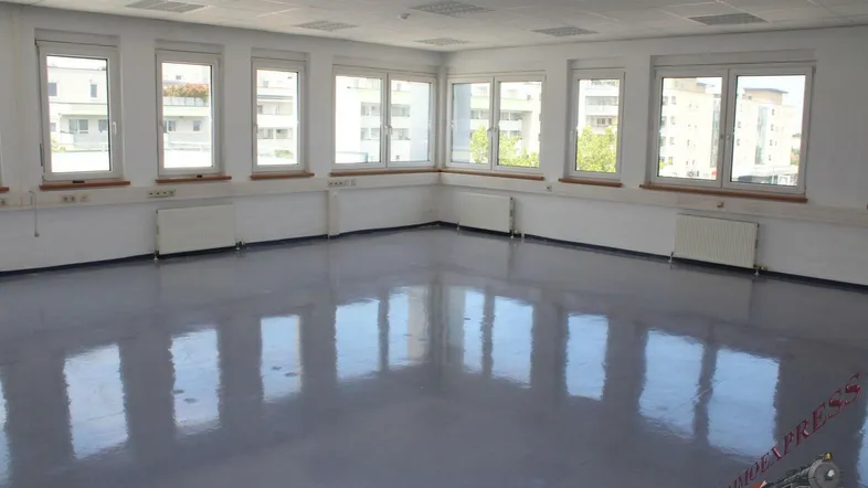 Heller, leerer Büroraum mit vielen Fenstern, grauem Vinylboden und weißen Wänden.