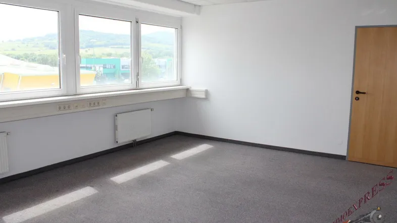 Leerer Büroraum mit großen Fenstern, Teppichboden und Blick auf die grüne Landschaft.