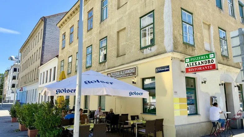 Eckgebäude mit einer Pizzeria im Erdgeschoss und Außenterrasse an einem sonnigen Tag in urbaner Umgebung.