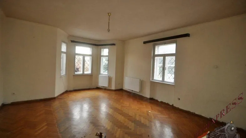 Helles Wohnzimmer mit Fischgrätparkett, Erkerfenstern und Heizkörpern, bereit zur Renovierung.