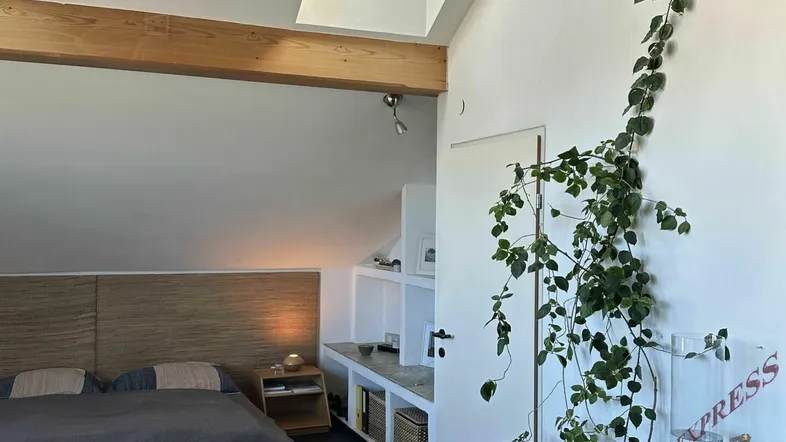 Helles Schlafzimmer mit Holzbalken, Dachfenster und einer Kletterpflanze, die dem Raum eine natürliche Note verleiht.