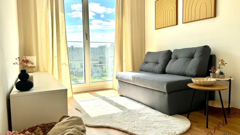 Helles Wohnzimmer mit grauem Sofa, Holzboden und großen Fenstern mit Blick ins Grüne.