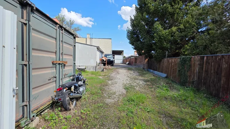 Schotterweg zwischen einem Container und einem Holzzaun, mit einem Motorrad am Rand.