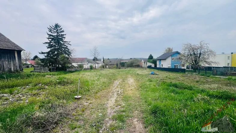 Ungenutztes Grundstück mit Schotterweg und umliegender Bebauung unter bewölktem Himmel.
