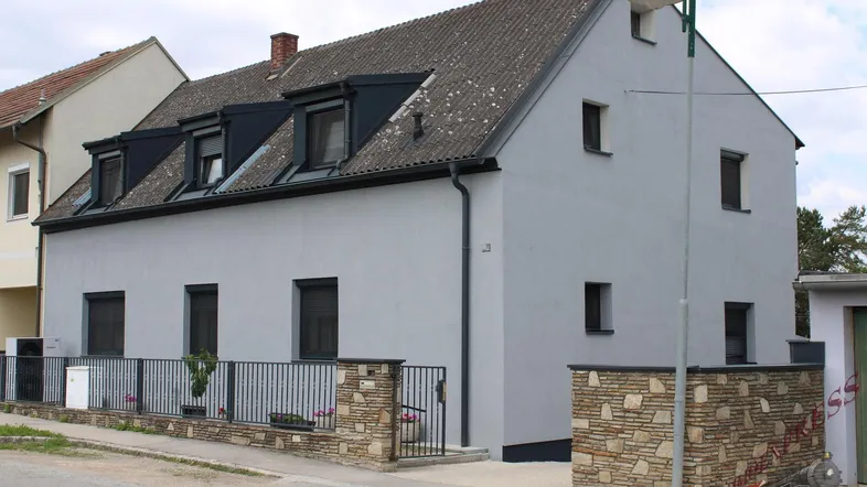 Graues Einfamilienhaus mit Satteldach, Gaubenfenstern und dekorativer Steinmauer am Eingangsbereich.