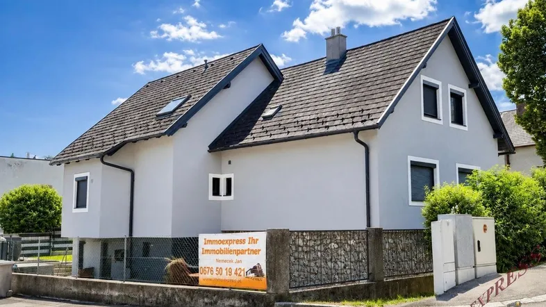Graues Einfamilienhaus mit Satteldach und Steinfassade im unteren Bereich unter blauem Himmel.