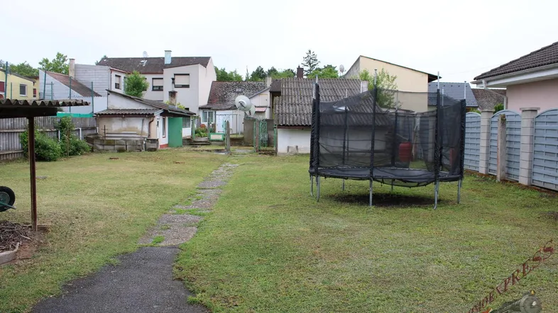 Großer Garten mit Rasenfläche, Trampolin und einem gepflasterten Weg, umgeben von Nachbargebäuden.