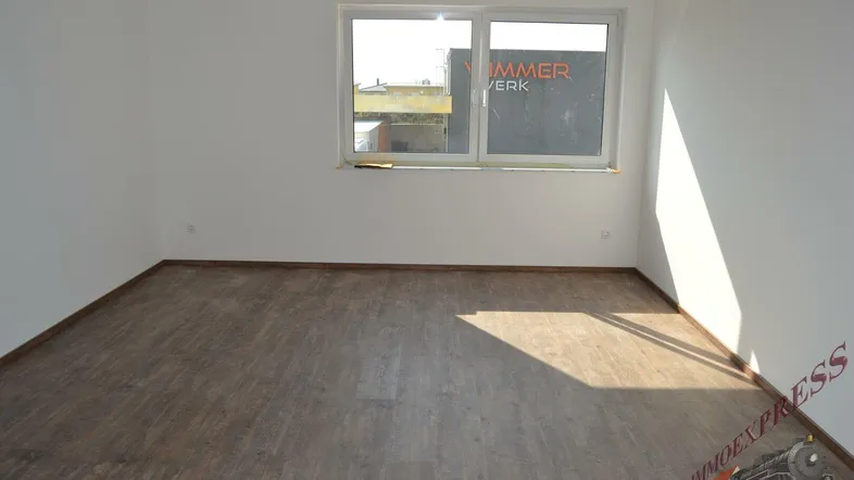 Leerer Raum mit Laminatboden und einem Fenster, das Tageslicht hereinlässt und eine Außenansicht zeigt.