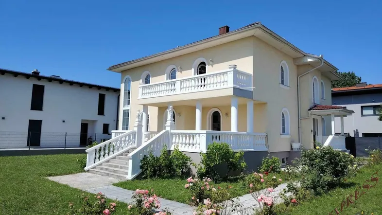 Elegante Villa mit klassischer Architektur, Balkon und gepflegtem Vorgarten unter blauem Himmel.