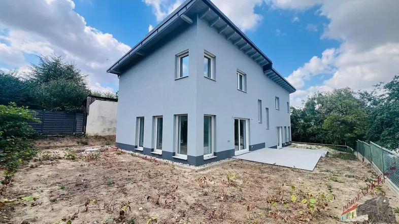 Neubau-Einfamilienhaus mit moderner grauer Fassade, großen Fenstern und einer vorbereiteten Terrasse.