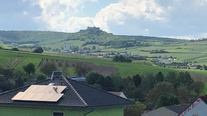 Blick auf die grüne Landschaft mit Weinbergen und einer Burg in der Ferne, im Vordergrund ein Haus mit Solaranlage.