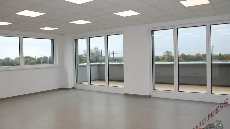 Heller, leerer Raum in einem Neubau mit großen Fenstern, Fliesenboden und Zugang zu einem Balkon. Die Decke ist mit integrierten Leuchten ausgestattet und bietet einen weiten Blick ins Grüne.