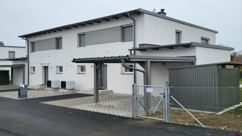 Modernes Reihenhaus mit Carport und gepflastertem Vorplatz unter bewölktem Himmel.