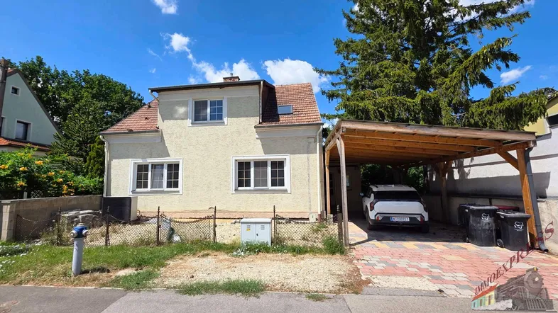 Freistehendes Haus mit heller Fassade, Satteldach und einem überdachten Carport auf gepflastertem Hof.