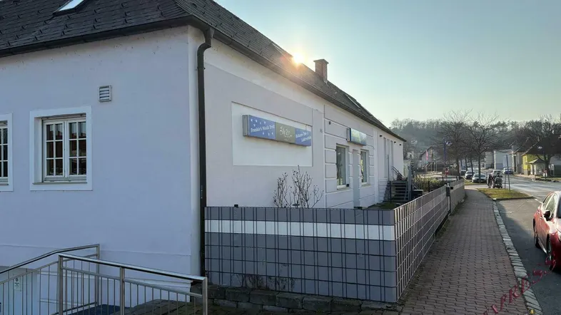 Gebäudeansicht von der Straße mit Sonnenschein und einem Zaun im Vordergrund.