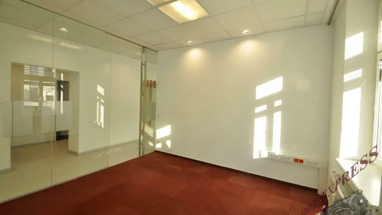 Helles Bürozimmer mit rotem Teppichboden, weißen Wänden und einer modernen Glasabtrennung zu einem weiteren Raum.