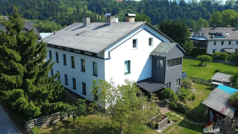 Hausansicht mit weißer Fassade, grauem Dach und umgebender grüner Landschaft.