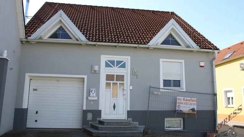 Graues Einfamilienhaus mit rotem Ziegeldach, weißer Haustür und Garagentor.