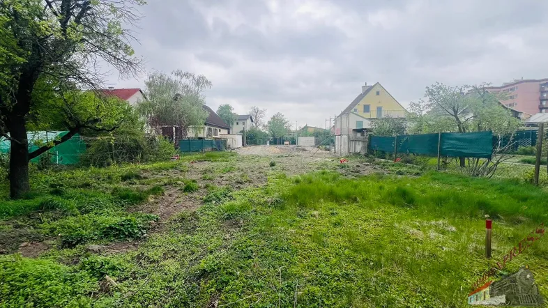 Unbebautes Grundstück mit Gras und Erde, umgeben von Zäunen und Häusern in der Ferne.