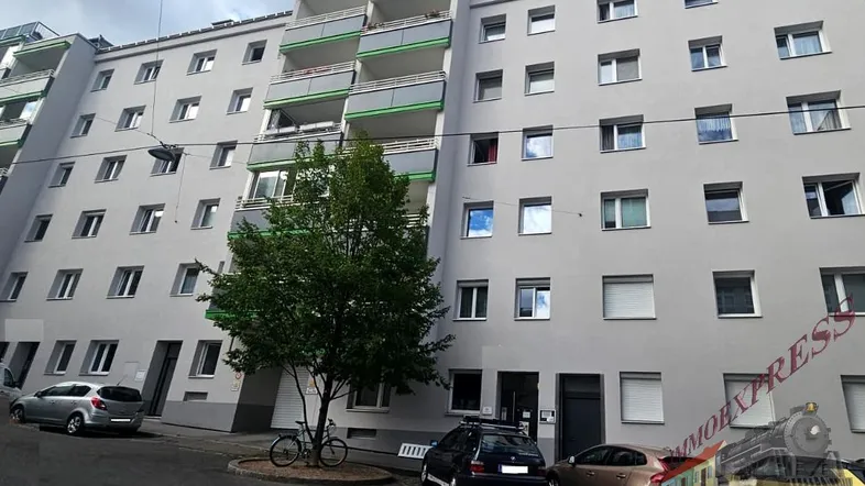 Mehrfamilienhaus mit grauer Fassade und grünen Balkonen in städtischer Umgebung.