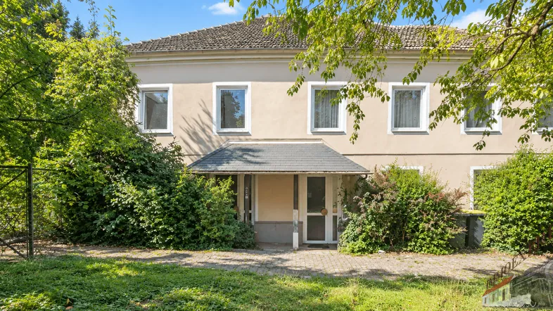 Fassade eines Hauses mit Eingangsbereich, umgeben von üppigem Grün und Bäumen.