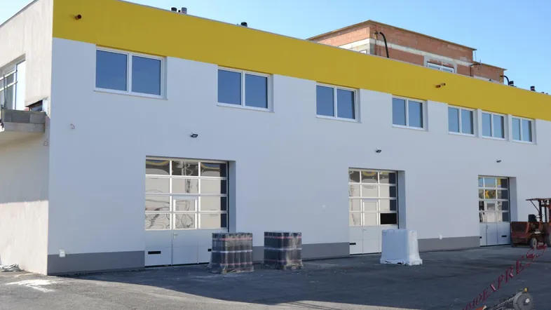 Modernes Geschäftsgebäude mit weißer Fassade, gelbem Akzentstreifen und mehreren großen Garagentoren, umgeben von einer asphaltierten Fläche.