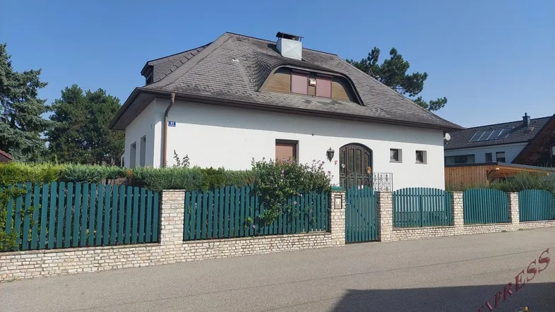 Weißes Einfamilienhaus mit Mansarddach und grünem Holzzaun an der Straße.