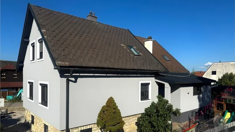 Graues Einfamilienhaus mit Satteldach und kleinen Fenstern unter blauem Himmel.
