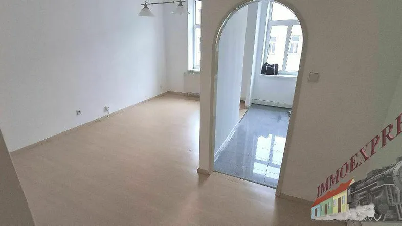 Helles Zimmer mit Laminatboden und weißen Wänden, durch einen Bogen mit einem weiteren Raum verbunden.