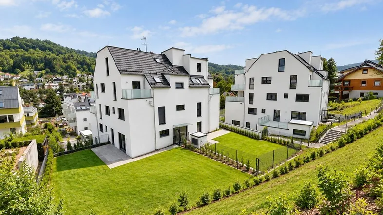 Moderne weiße Mehrfamilienhäuser mit Gärten und Blick auf die grüne Hügellandschaft.