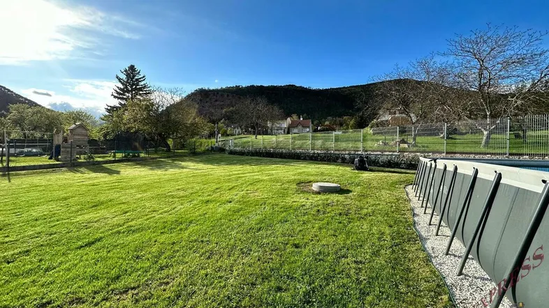 Großer grüner Garten mit Rasenfläche, Aufstellpool und Spielgeräten vor einer malerischen Hügellandschaft.